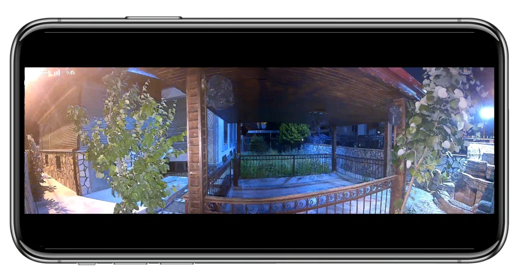 A wide-angle view from a security camera shows a covered porch with metal railings, greenery, and a small garden outside at night.